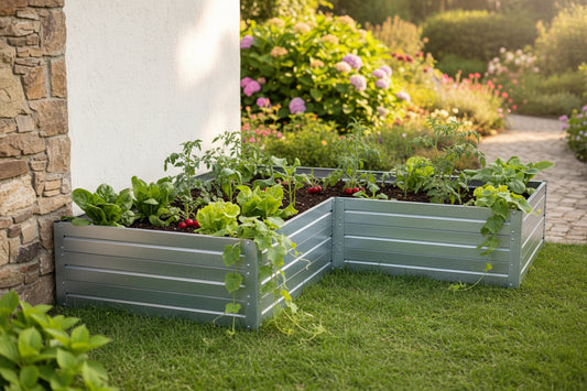 Two L-shaped raised garden beds made of horizontal slats in a muted sage green, filled with leafy greens and herbs, set on a green lawn next to a white wall and stone pillar in a sunny garden.